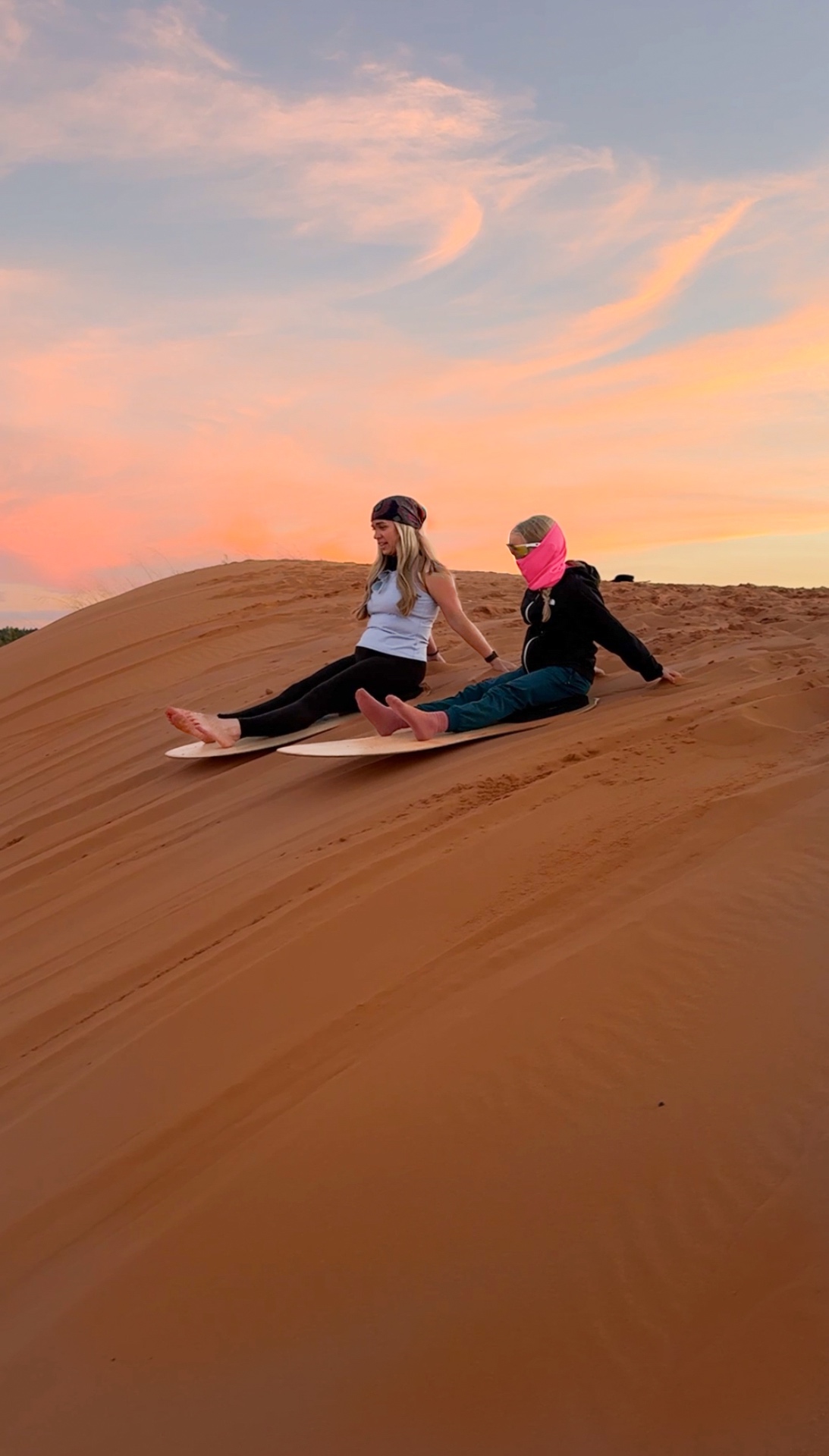 Brittany and Aniston sitting on sandboards at the top of a pink dune as the sky turns pink and orange at sunset