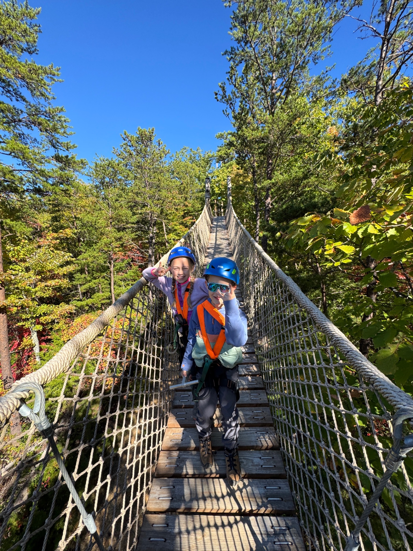 Aniston and Noah giving thumbs up while crossing a rope skybridge in the trees