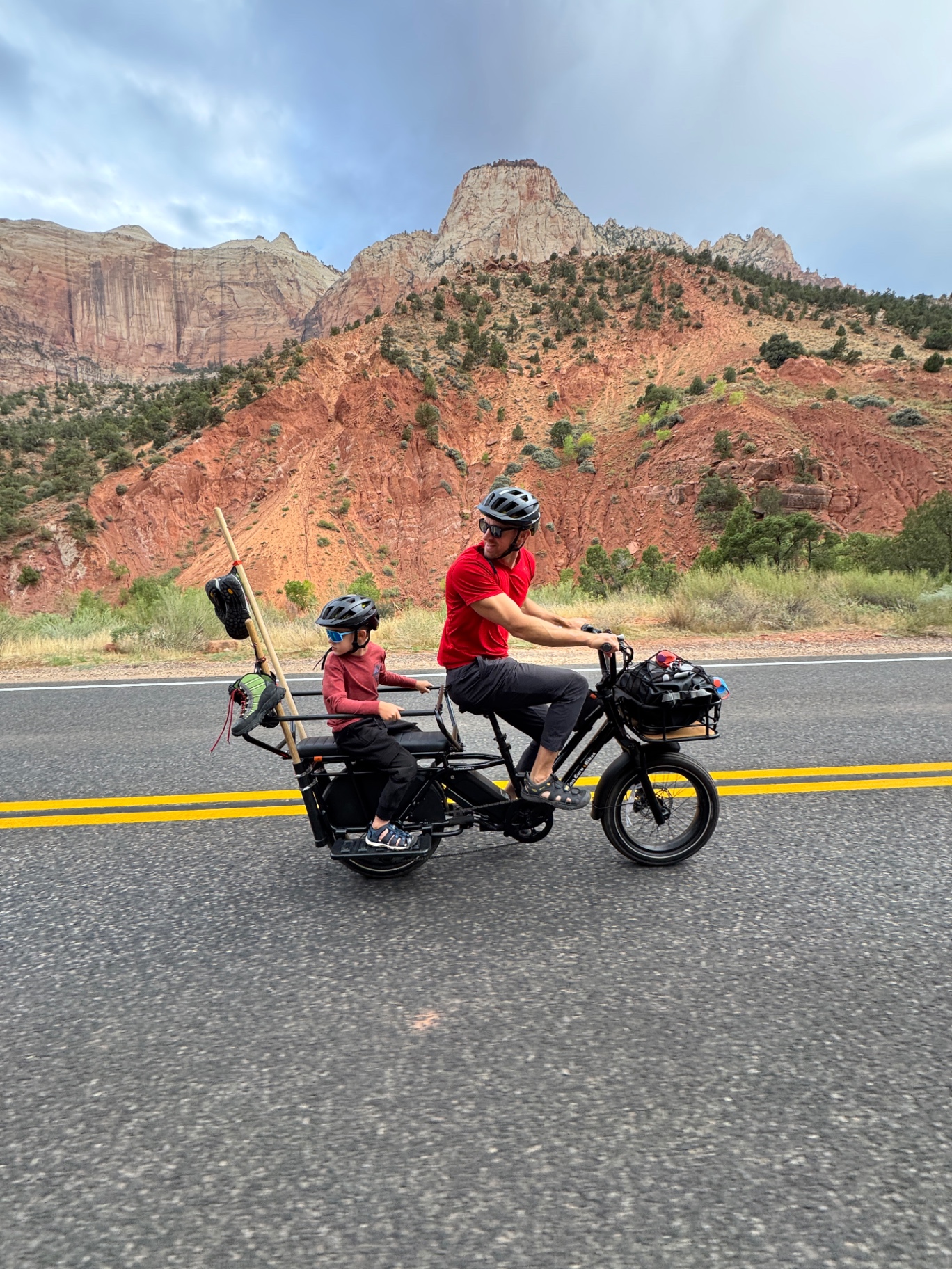 Jake riding an e-bike with Noah on the back through Zion National Park with red rock cliffs in the background|IMG:/__l5e/assets-v1/c7ebf42b-31b3-4436-a1c5-7ae5a3b596ff/img_2352.jpeg|Brittany on her e-bike waving back at the camera on the empty Zion scenic drive with canyon walls behind her