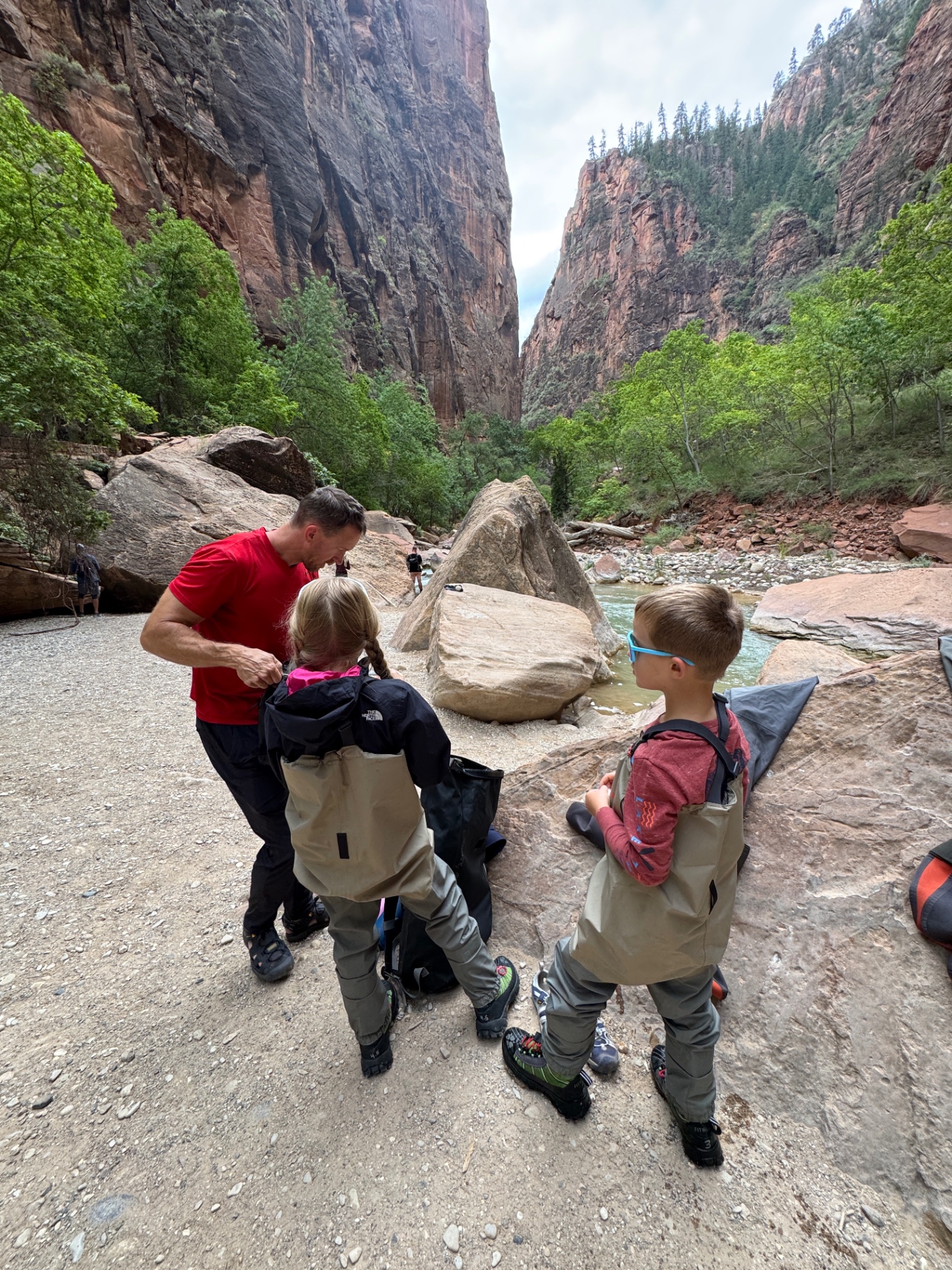 Jake helping Aniston and Noah zip into their waders at the trailhead before hiking The Narrows in Zion|IMG:/__l5e/assets-v1/610b30e5-f40b-4106-ba7b-4a8705022937/img_2362.jpeg|Jake biking with Noah on the back through Zion, hiking boots tied to a pole strapped to the cargo bike