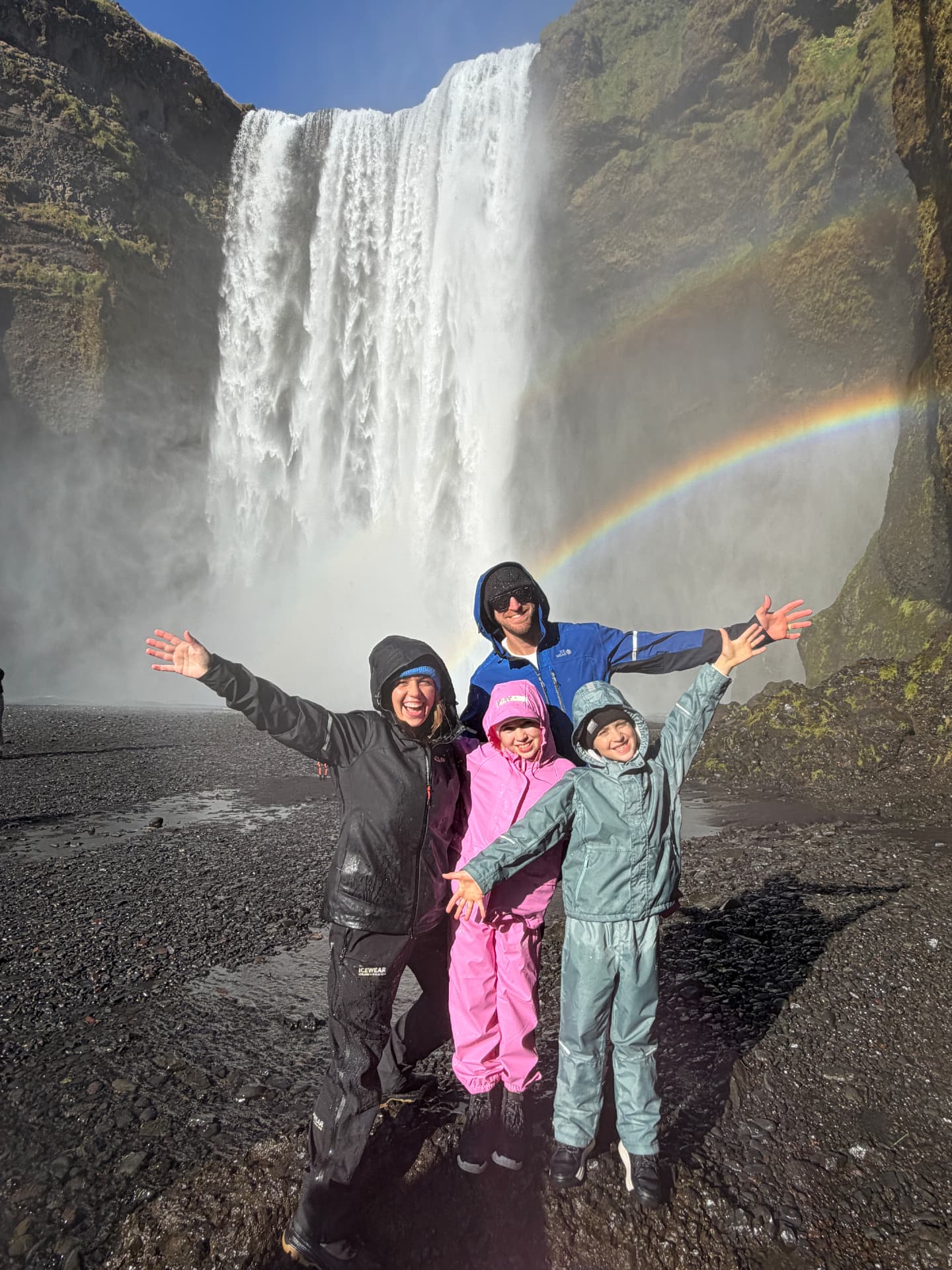 All four of us in our rain gear in front of a waterfall with a rainbow on the drive to Vík