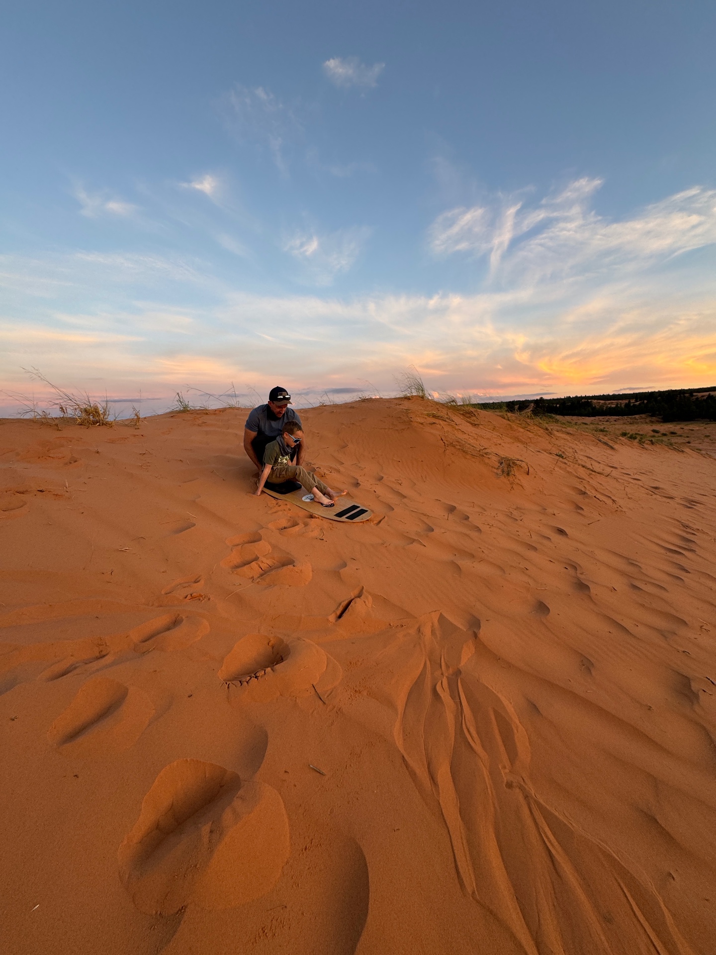 Jake and Noah sitting together at the top of a dune with a sandboard, sunset sky behind them