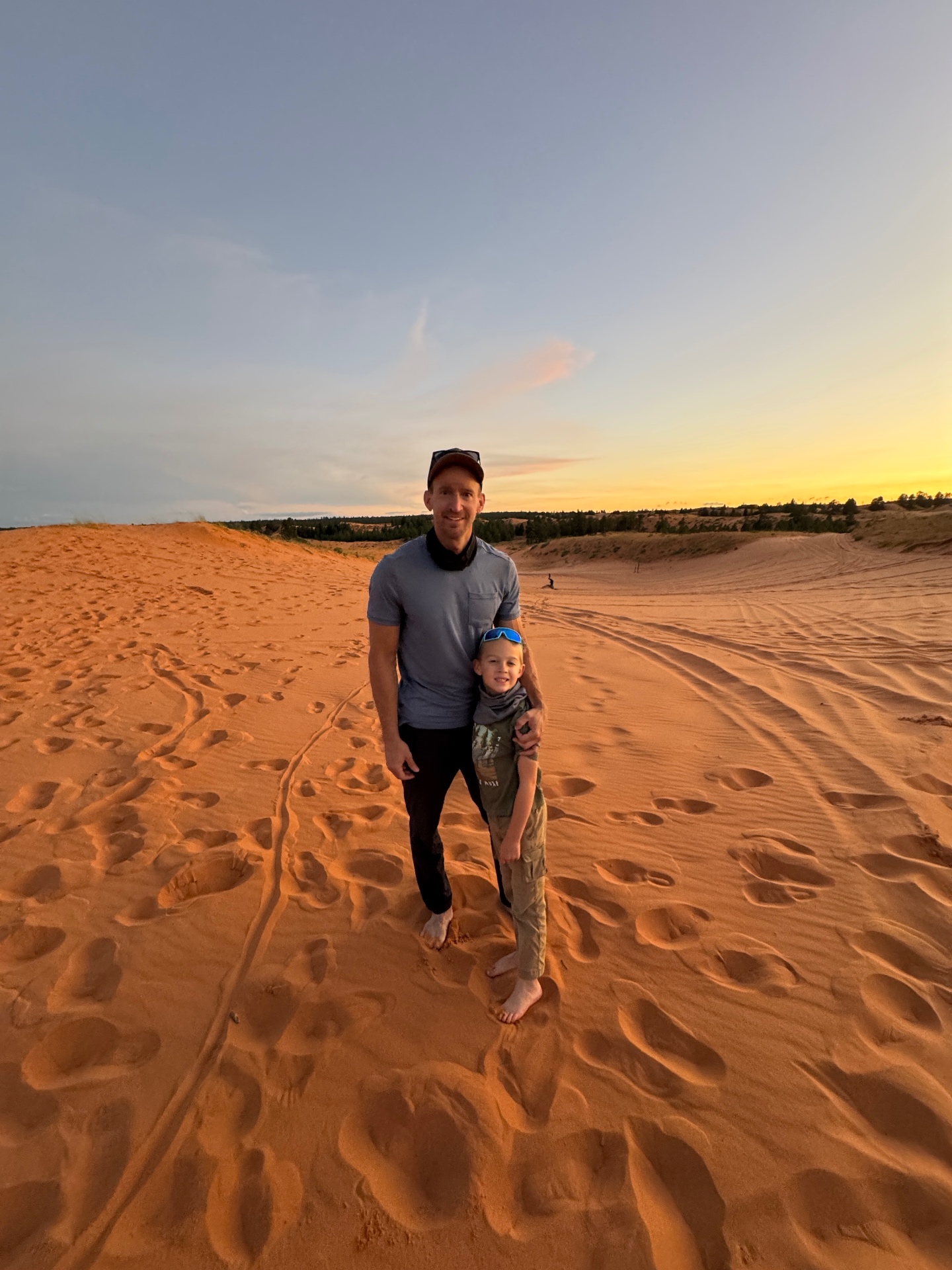 Jake and Noah standing barefoot in the pink sand with the sun setting over the dunes behind them