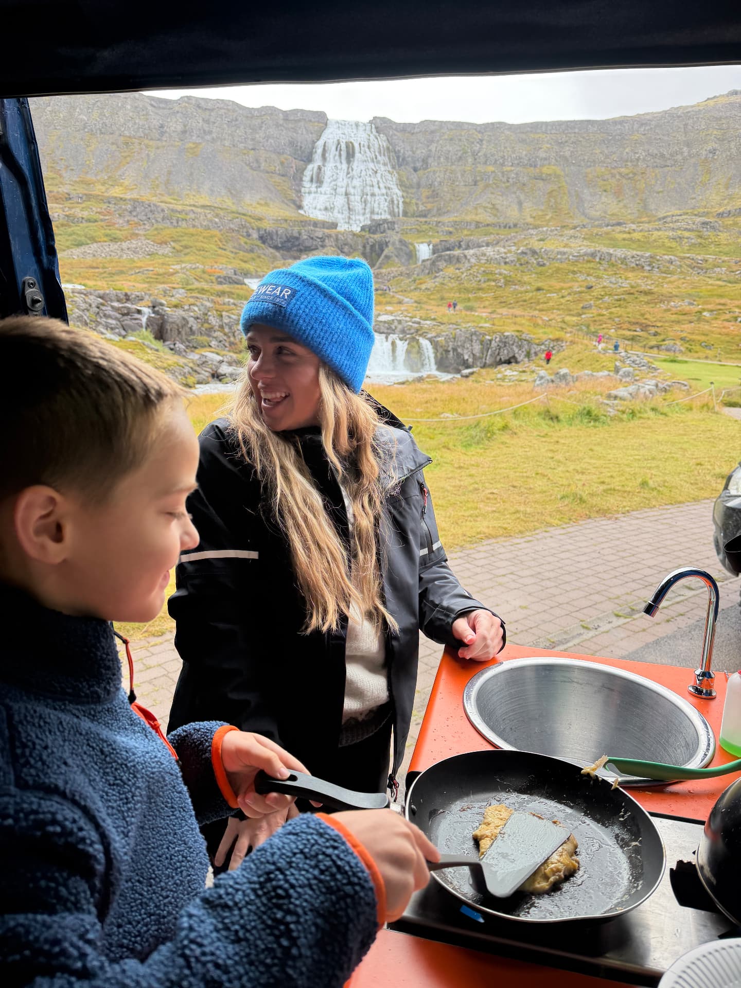 Brittany and one of the kids making food in the campervan with a waterfall visible through the back doors