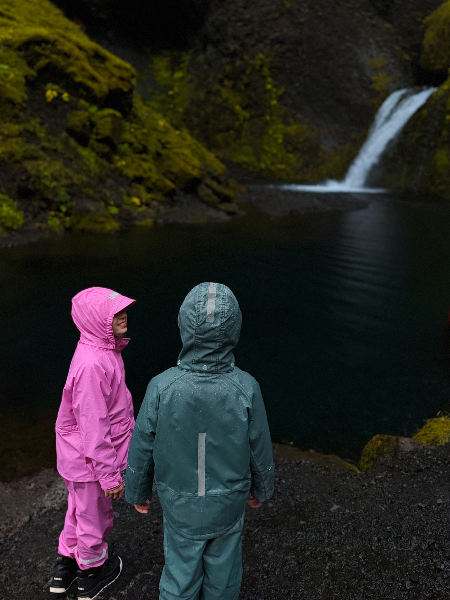 Aniston and Noah in their rain gear at a mossy waterfall, proof that the right shells make rainy days fun