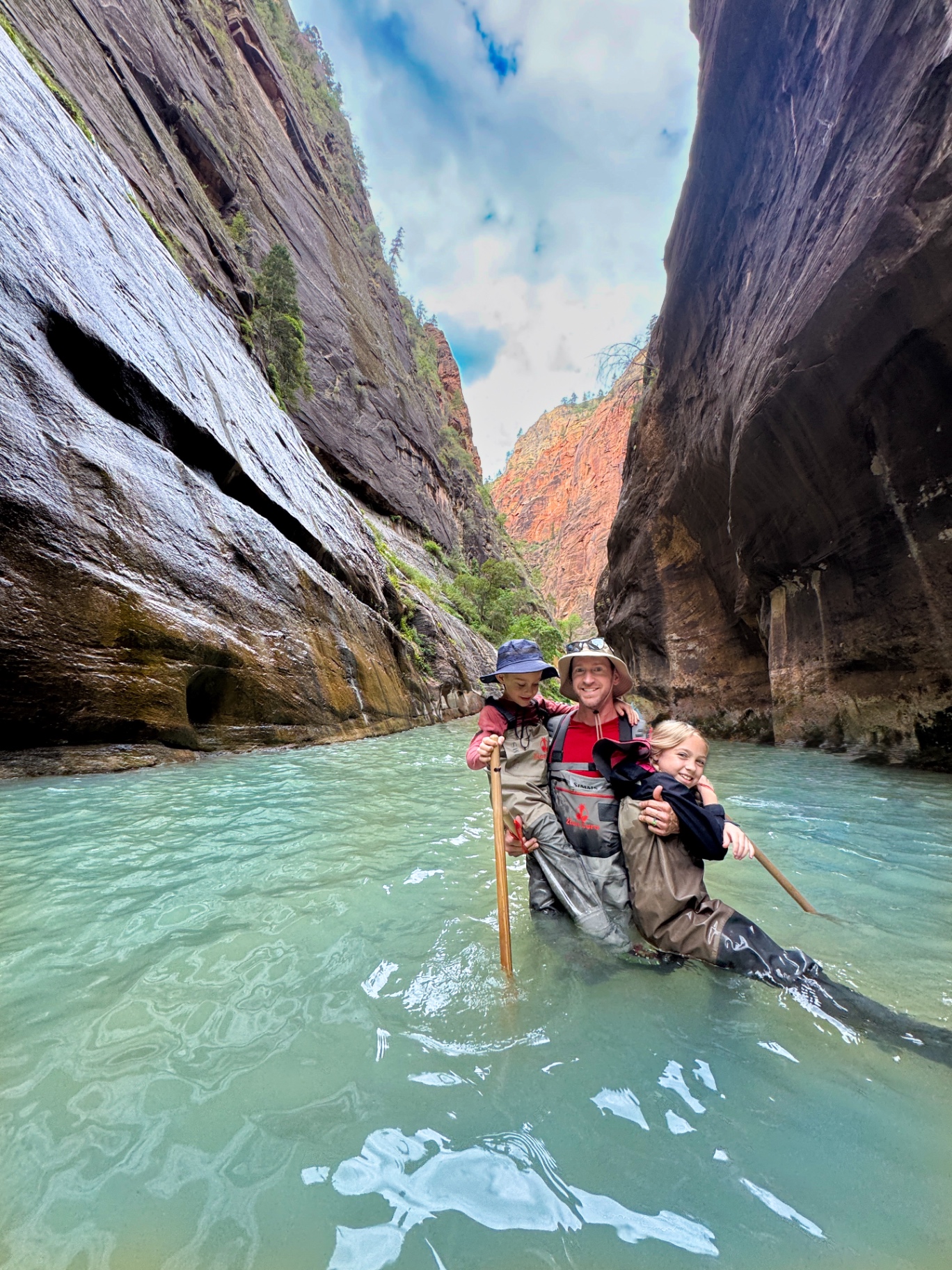Jake holding Aniston and Noah in waders standing chest-deep in the turquoise Virgin River with the towering walls of The Narrows rising above them|IMG:/__l5e/assets-v1/dcb6ef33-2c61-4cec-b86d-25e67052ca6e/img_2447.jpeg|Aniston and Noah sitting on a red rock ledge in their waders with hiking poles inside The Narrows in Zion