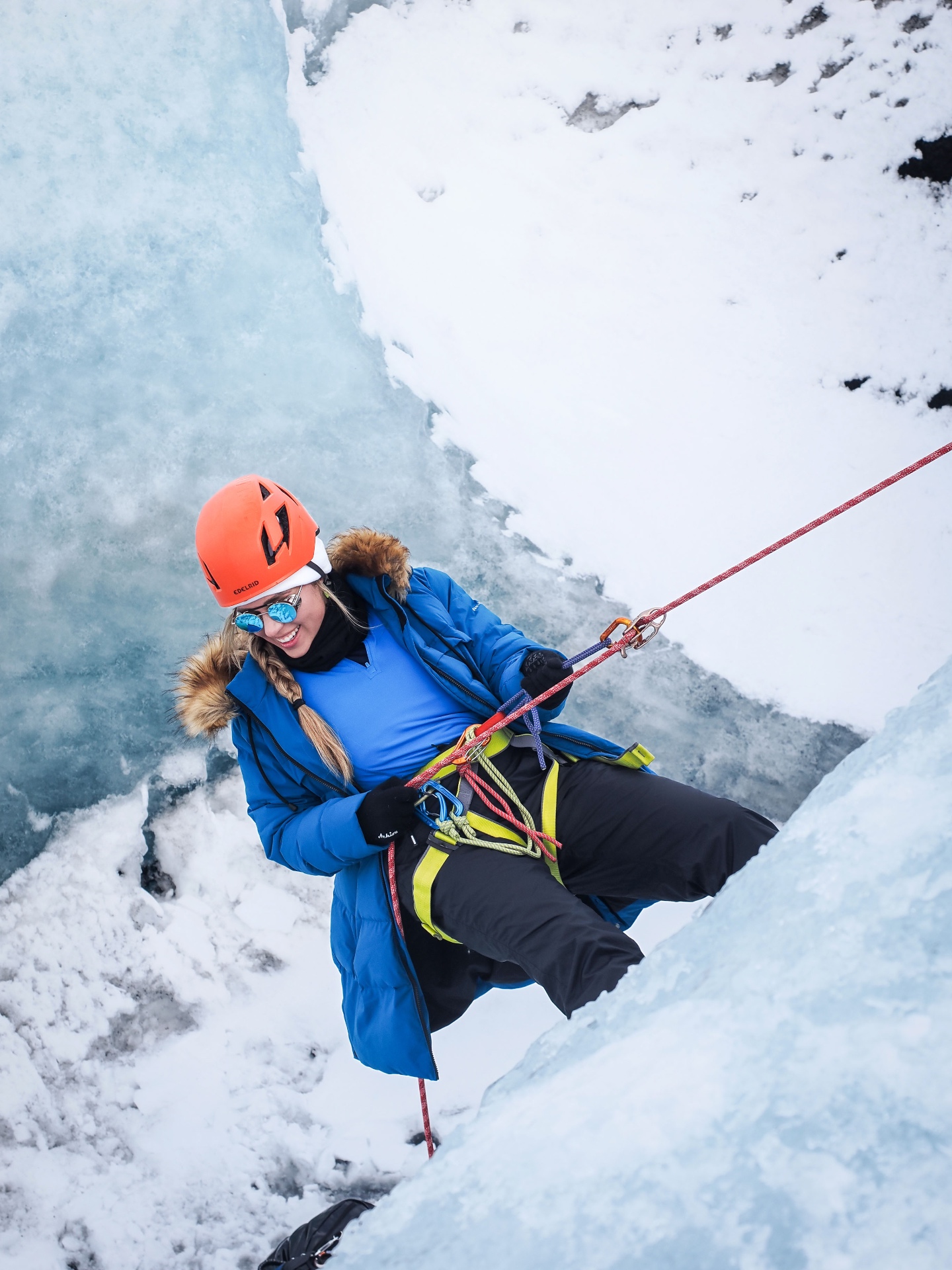 Brittany rappelling down a glacier wall in a blue jacket and orange helmet