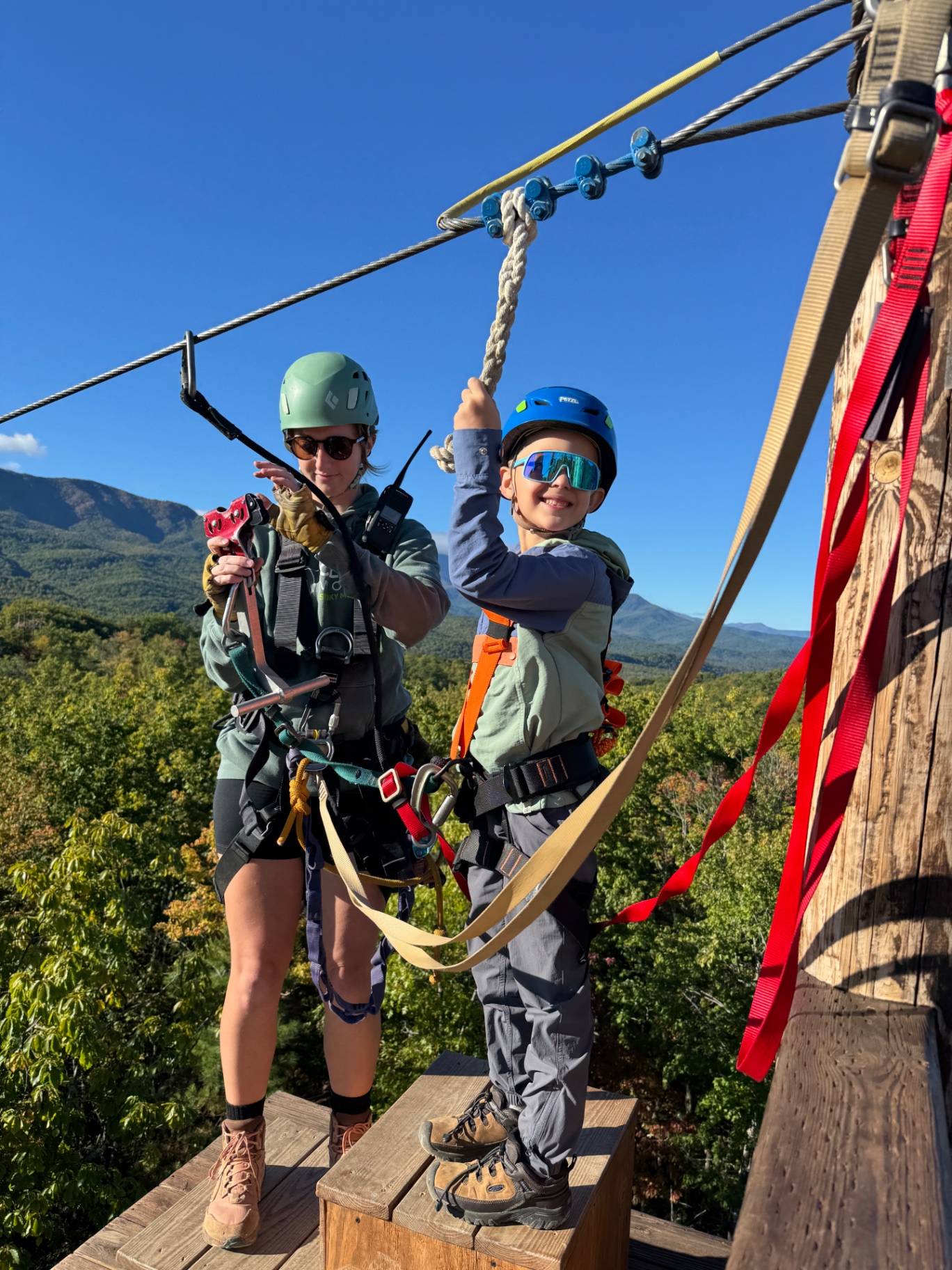 Brittany clipping Noah into the side-by-side zipline platform with the Smoky Mountains behind them