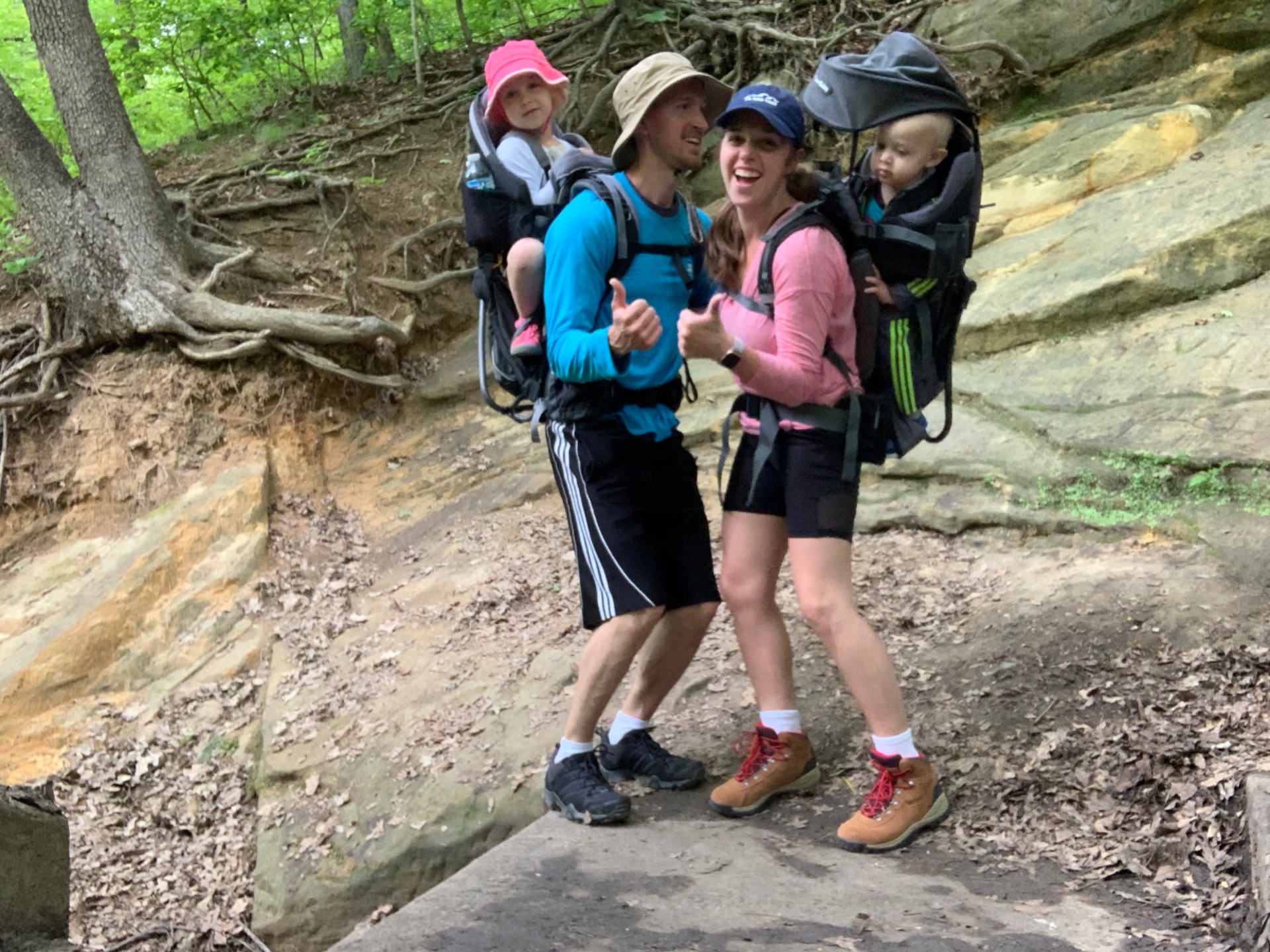 Jake and Brittany hiking with Aniston and Noah in backpack carriers, giving thumbs up on the trail