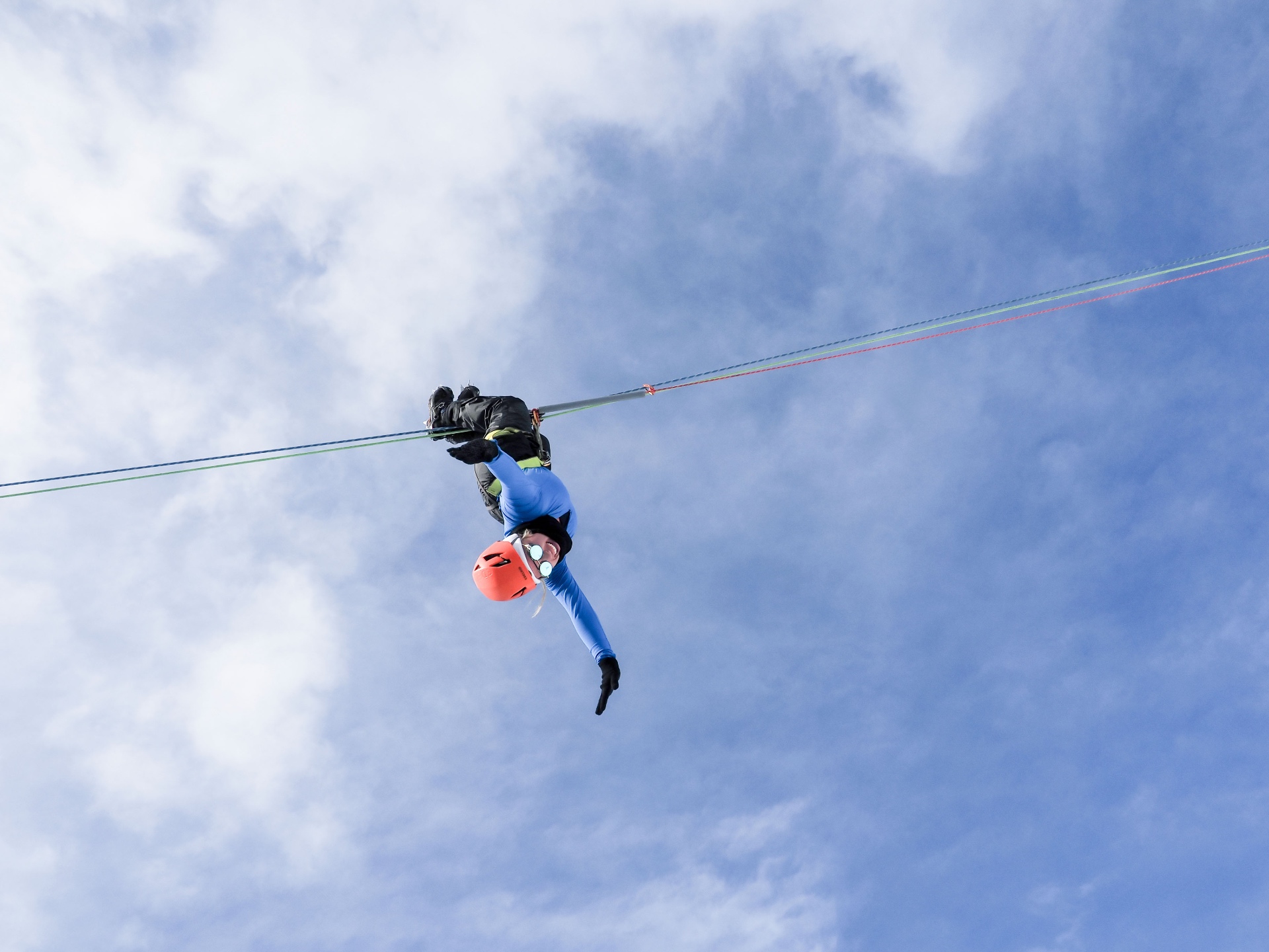 Brittany hanging upside down from the glacier zipline against a bright cloudy sky