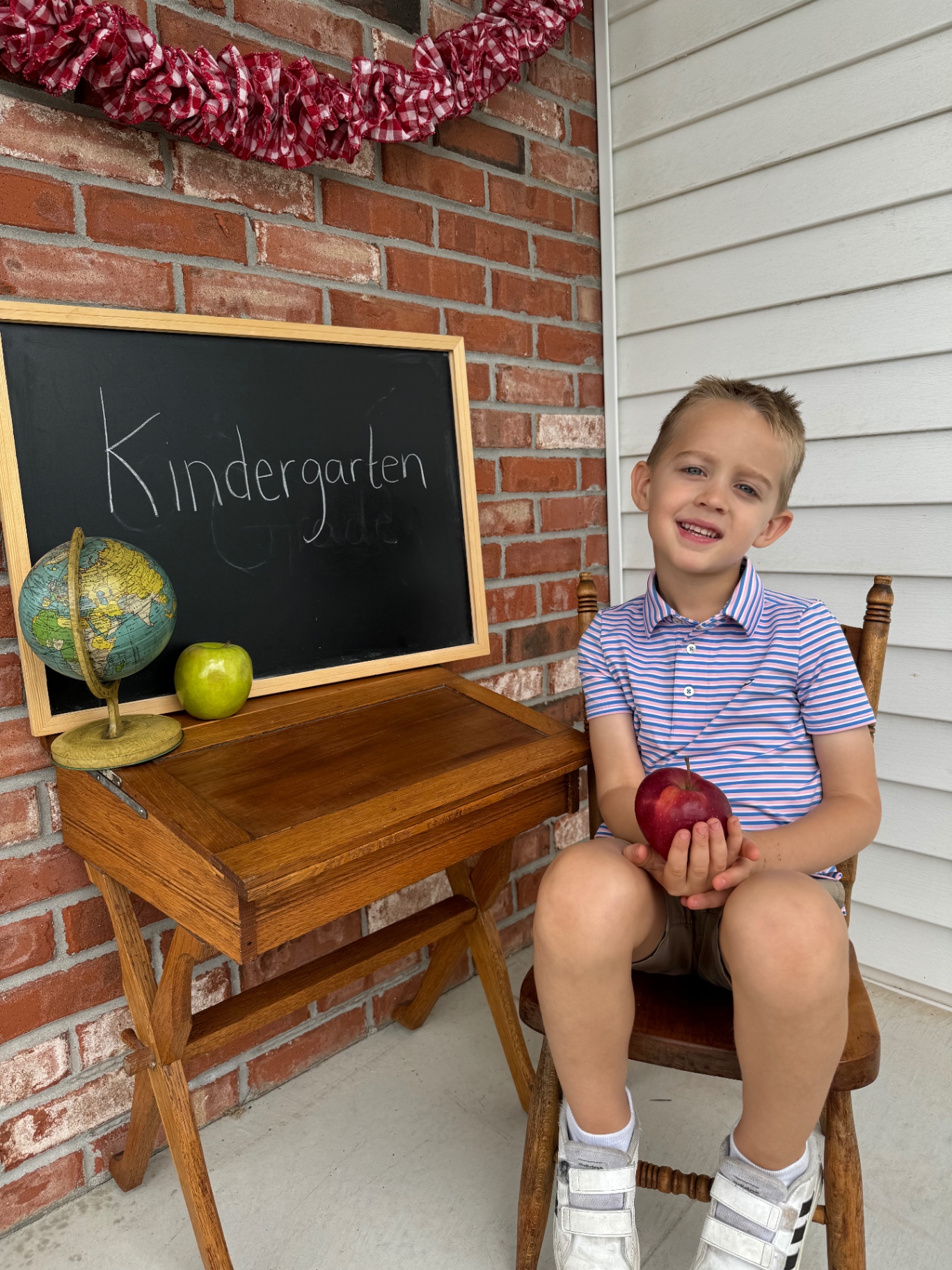 Noah holding an apple next to a chalkboard that reads Kindergarten on his first day of homeschool