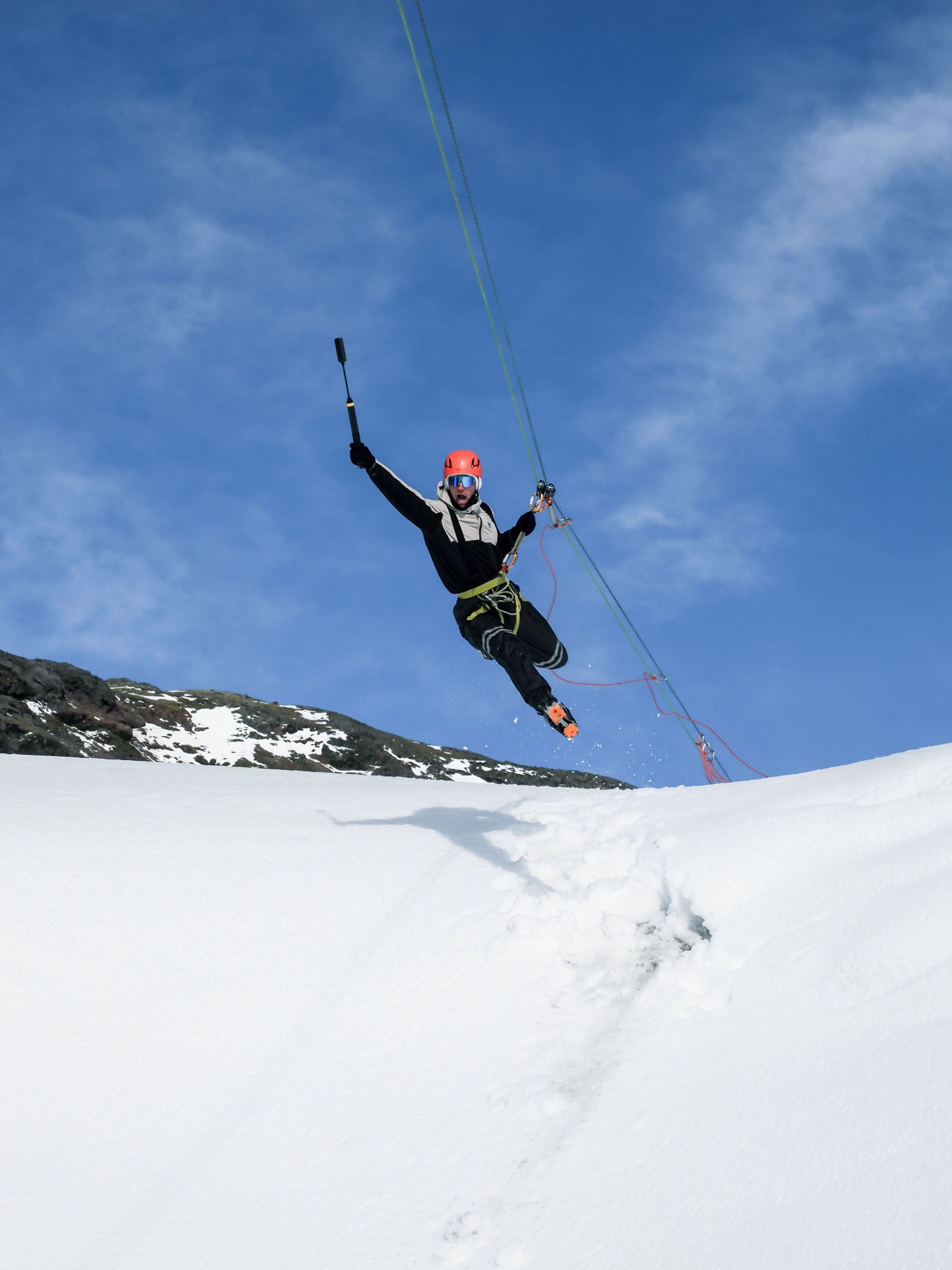 Jake mid-air off the glacier on a rope, holding a GoPro, bluebird sky behind him