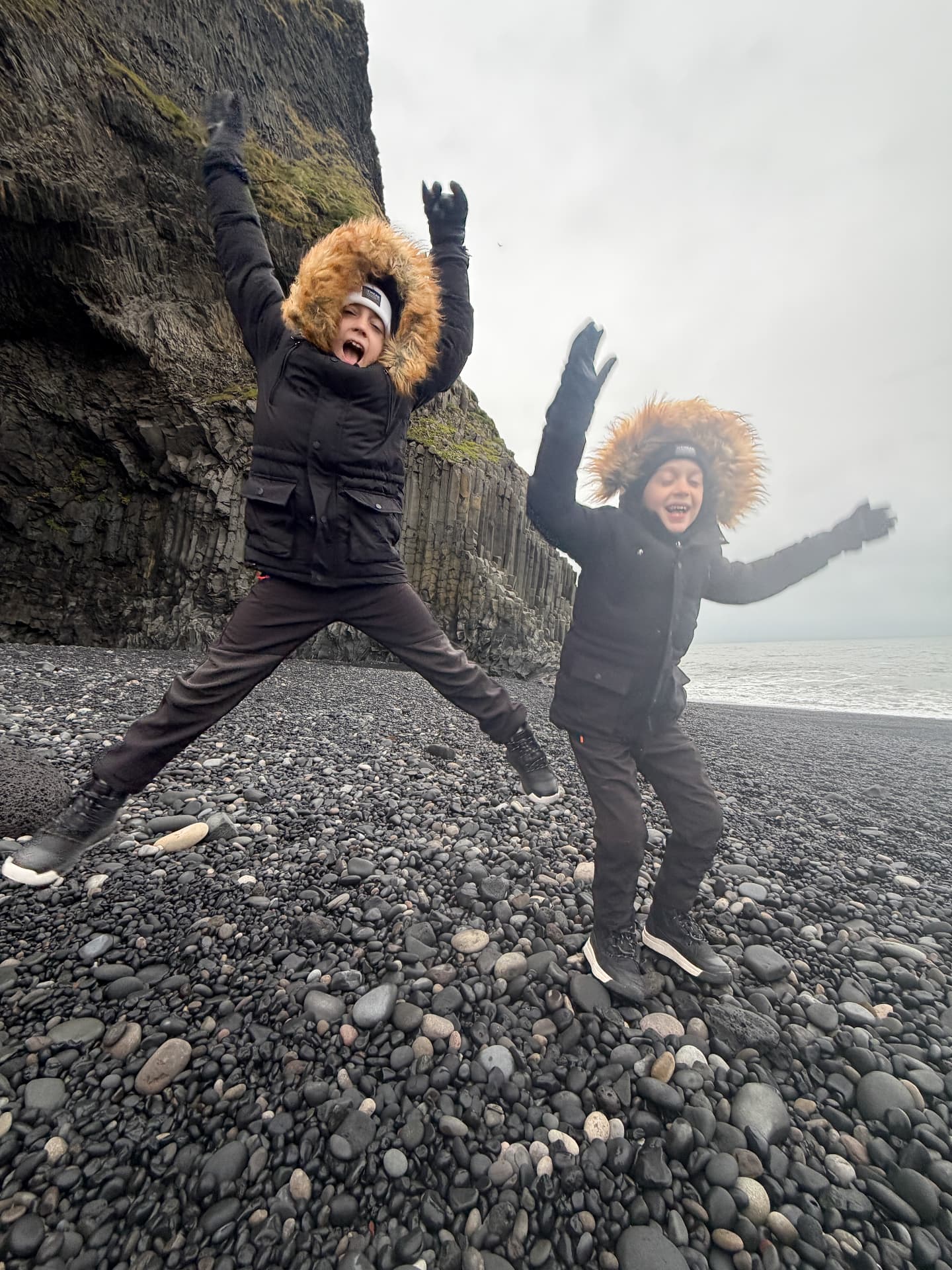 Aniston and Noah jumping on the black sand beach near Vík with the basalt cliffs behind them