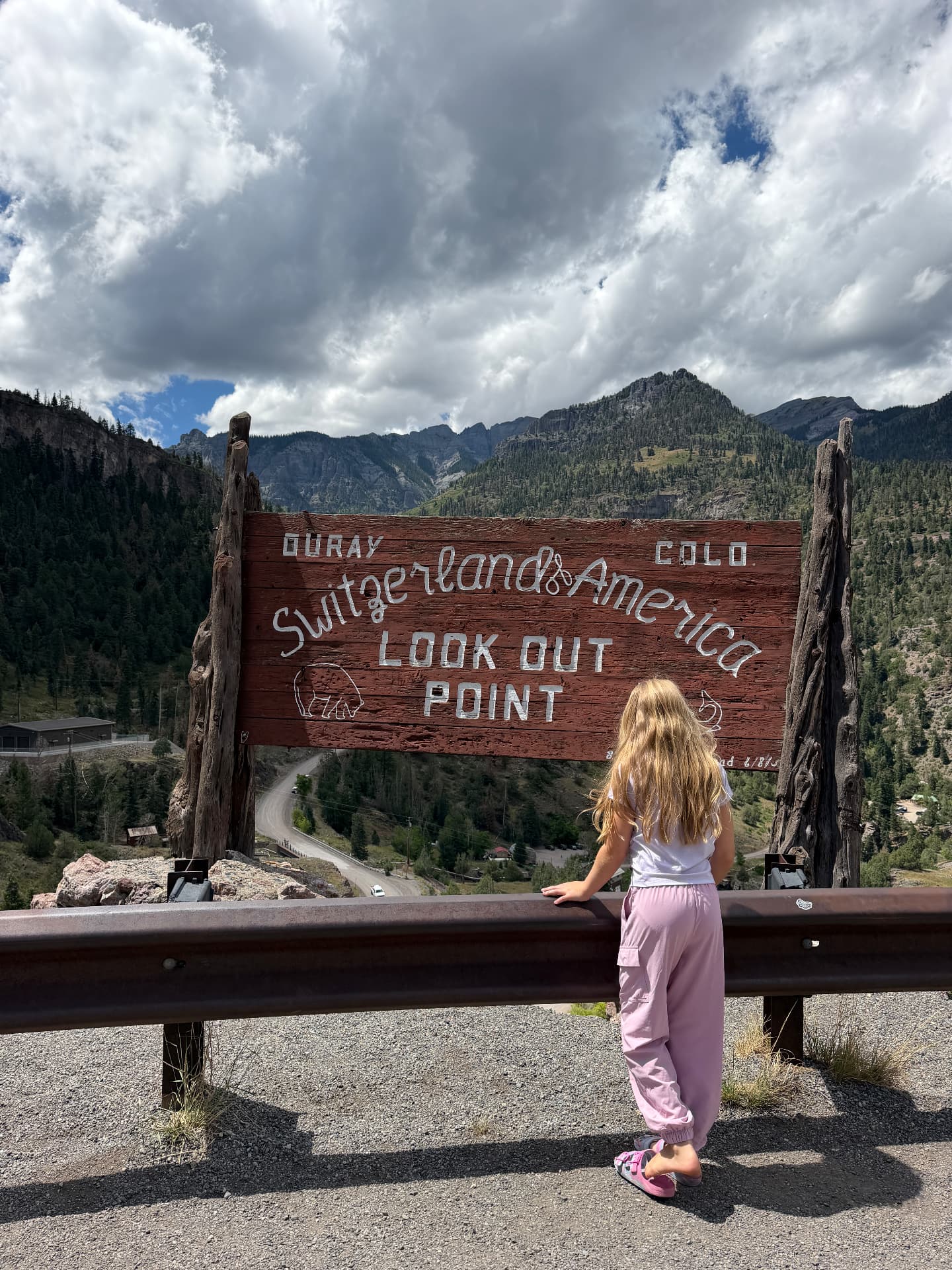 A young girl looking out at the Switzerland of America Lookout Point sign near Ouray, Colorado with the San Juan Mountains behind